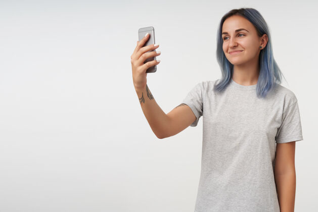智能手机室内肖像的年轻短发积极纹身女性保持手机在举起的手和微笑愉快 隔离在白色室内化妆女性