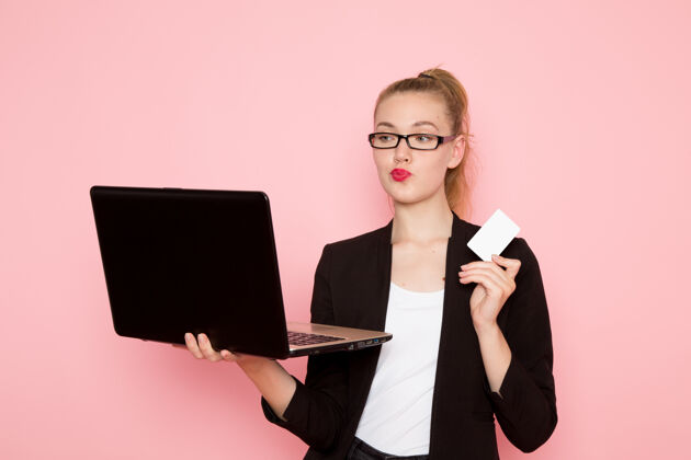 公司身穿黑色夹克的女上班族正面图 手持卡片 在粉色墙上使用笔记本电脑工人秘书商务女性