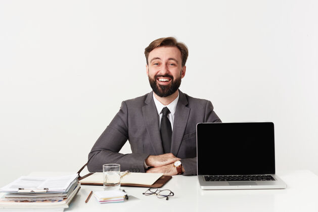 领带快乐的年轻漂亮的胡须男子穿着灰色西装 打着领带 拿着现代化的笔记本电脑和笔记本在办公室工作 双手合十放在桌子上 面带微笑地看着前方穿发型未剃