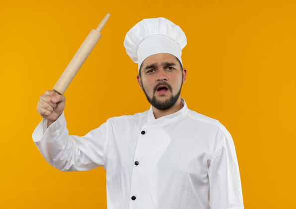 厨师穿着厨师制服的年轻男厨师举起擀面杖孤立在橙色的空间橙色年轻制服
