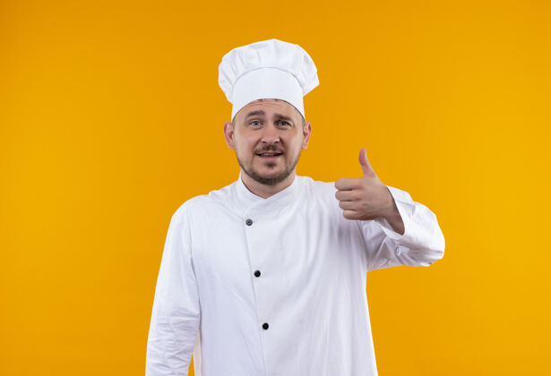 烹饪身着厨师制服 面带微笑的年轻帅哥厨师在孤立的橙色空间竖起大拇指厨师秀橙色