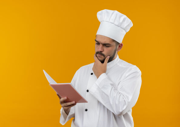 男性体贴的年轻男厨师穿着厨师制服 手放在下巴上 手拿着便签本看着橙色的空间年轻厨师制服