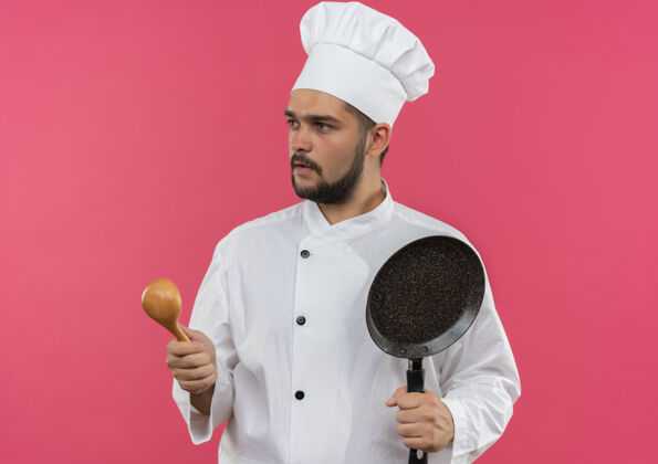 平底锅年轻的男厨师穿着厨师制服 拿着勺子和煎锅 看着旁边孤立的粉色空间持有侧煎炸