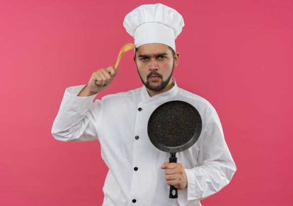 未出租穿着厨师制服的年轻男厨师拿着煎锅 用勺子摸着头 被隔离在粉红色的空间里男性持有厨师