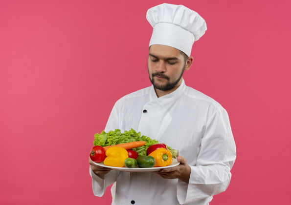 厨师年轻的男厨师穿着厨师制服 拿着盘子看着隔离在粉红色空间里的蔬菜空间年轻持有