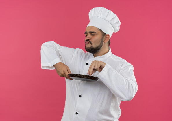 制服穿着厨师制服的年轻男厨师手拿煎锅 手指放在上面 闭着眼睛 孤立地站在粉红色的空间里年轻薯条放