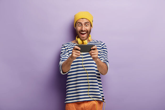 人有趣开朗的男性玩家通过智能手机玩电子游戏 戴着黄色帽子和条纹套头衫 沉迷于现代科技 孤立在紫色的墙上 查看新的应用程序玩学生积极