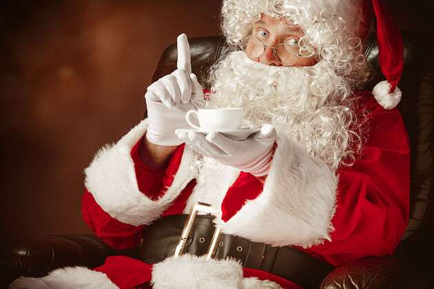杯子一个穿着圣诞老人服装 留着奢华的白胡子 戴着圣诞老人的帽子 穿着红色服装的男人坐在椅子上喝着咖啡胡子人旧的