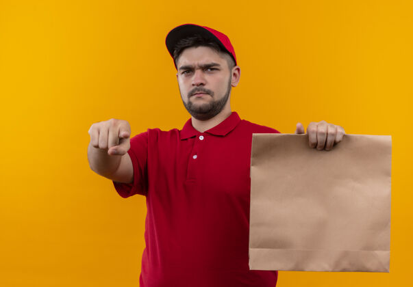 制服年轻的送货员身穿红色制服 戴着帽子 手里拿着纸包 用手指指着镜头 满脸怒容红色站着纸
