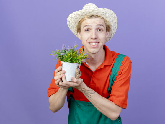 年轻年轻的园丁 穿着连身衣 戴着帽子 手里拿着盆栽植物 站在紫色的背景上 面带微笑地看着镜头站着快乐看
