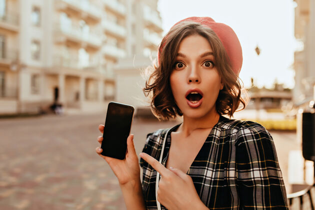 短裤在街上拿着智能手机的优雅女人惊呆了穿着红色贝雷帽的情绪激动的女孩的户外照片帽子快乐户外