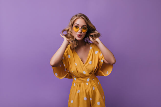 黄色可爱优雅的女孩戴着时髦的太阳镜抚摸着她的卷发优雅的金发女模特站在明亮的紫色上优雅模特金发