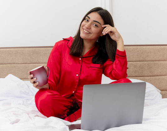 室内穿着红色睡衣的年轻美女坐在床上 拿着笔记本电脑和一杯咖啡 面带微笑地看着相机 在卧室的室内灯光背景下快乐而积极年轻笔记本电脑脸
