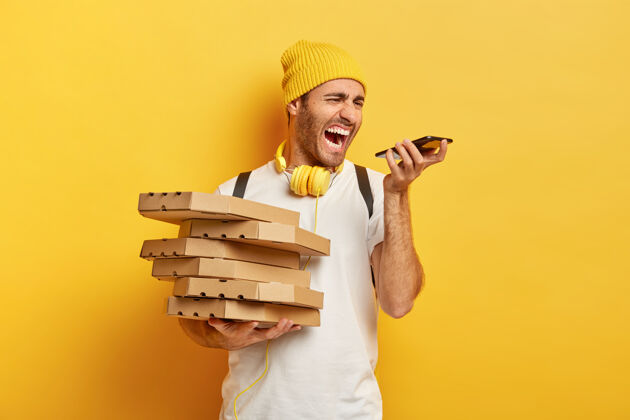 食物恼怒的披萨男快递员对着智能手机愤怒地尖叫 与客户进行恼人的对话 拿着一堆纸箱 戴着帽子和白色t恤 隔离在黄色的墙上快递人订单