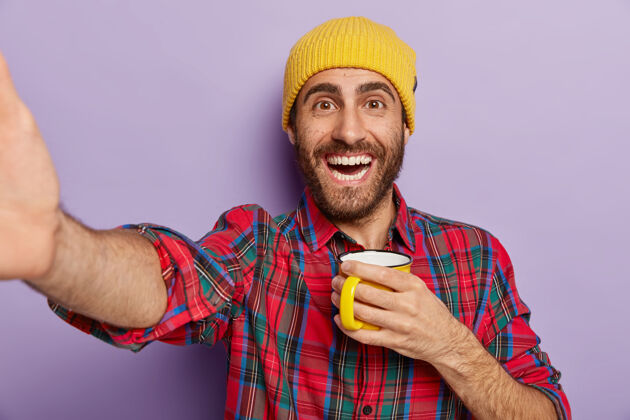 休闲快乐的白人男子在室内自拍 拿着咖啡或茶的杯子 享受休息和自由时间 戴着时尚的黄色帽子和格子衬衫隔离在紫色的墙上人和生活方式饮酒咖啡高兴