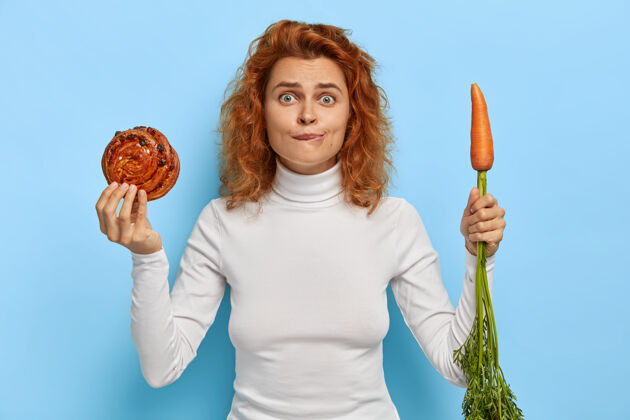 选择人 营养 节食和垃圾食品的概念尴尬的红发女人拿着新鲜可口的面包和胡萝卜 在蔬菜和糖果之间选择 穿着白色高领毛衣 站在室内发髻发型新鲜
