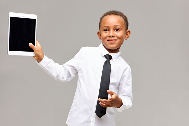 使用开朗可爱的非裔美国男孩穿着衬衫 打着领带 微笑着愉快地使用电子设备玩游戏或看动画片 手持数字平板电脑与空白显示的文字空间肖像可爱年轻