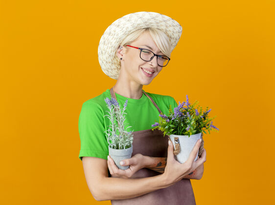 头发一位年轻的园丁 围裙上留着短发 戴着帽子 手里拿着盆栽植物 站在橙色的背景上 开心而积极地微笑着微笑年轻植物