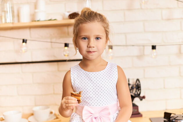饼干快乐的童年 有趣和快乐的概念室内拍摄的甜美可爱的女婴穿着漂亮的裙子坐在餐桌上 在时尚的厨房内部 笑着 嚼着美味的饼干或馅饼小年轻饼干