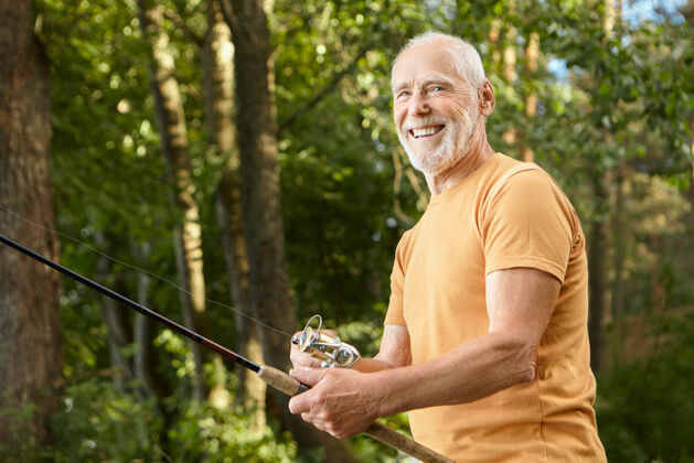 成熟健康微笑胡须的白人男性退休老人的肖像 身穿t恤 在户外与绿树握着鱼竿 享受垂钓休闲 休闲和自然的概念老人湖泊放松