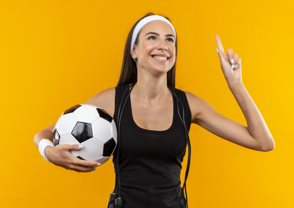 穿带着微笑的年轻漂亮的运动女孩戴着头带和腕带拿着足球 脖子上系着跳绳孤立在橙色的空间里头带足球周围