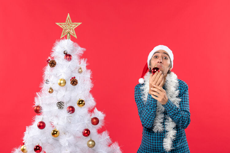 站着一个戴着圣诞老人帽子 穿着蓝色条纹衬衫 拿着装饰品的悲伤 不开心 情绪激动的年轻人男人树帽子