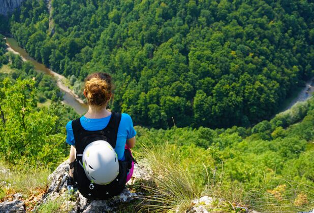 人一个女徒步旅行者坐在悬崖边俯瞰森林的高角度镜头女性河流岩石