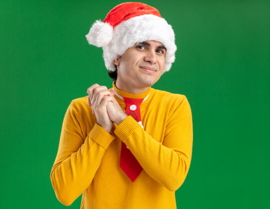 高领毛衣穿着黄色高领毛衣 戴着圣诞帽 打着滑稽领带的年轻人手牵着手 快乐地站在绿色的背景上等待惊喜在一起领带高兴