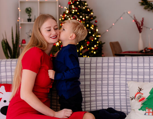 圣诞快乐在一间装饰有圣诞树的房间里 穿着红色连衣裙的快乐妈妈和坐在沙发上的小孩在亲吻快乐的妈妈小衣服装饰