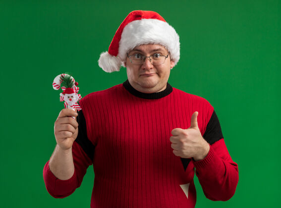 手杖高兴的成年男子戴着眼镜和圣诞老人帽子拿着糖果藤条饰物 竖起大拇指看着隔离在绿色背景上的相机展示圣诞快乐糖果