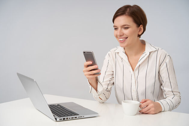条纹年轻快乐的棕色头发女性 自然妆容 手举手机 面带微笑 看着屏幕 在白色的茶杯上摆姿势桌子正式优雅
