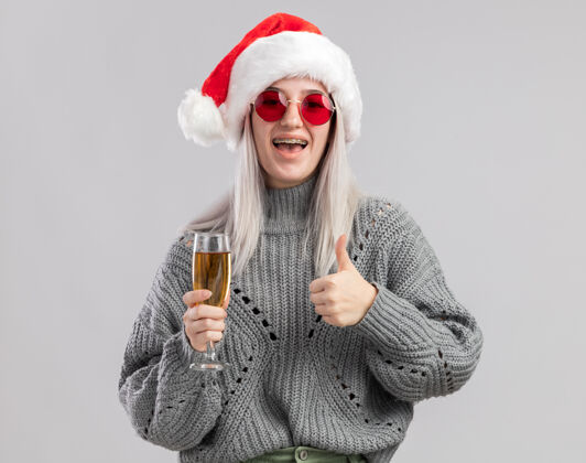女人年轻的金发女郎穿着冬衣 戴着圣诞帽 手里拿着一杯香槟 竖起大拇指 快乐而积极帽子年轻圣诞快乐