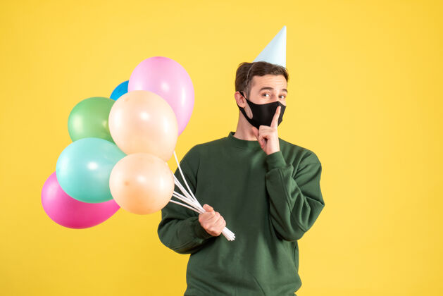 庆祝正面图：戴着派对帽的年轻人和五颜六色的气球站在黄色的墙上做嘘标志吹生日制造
