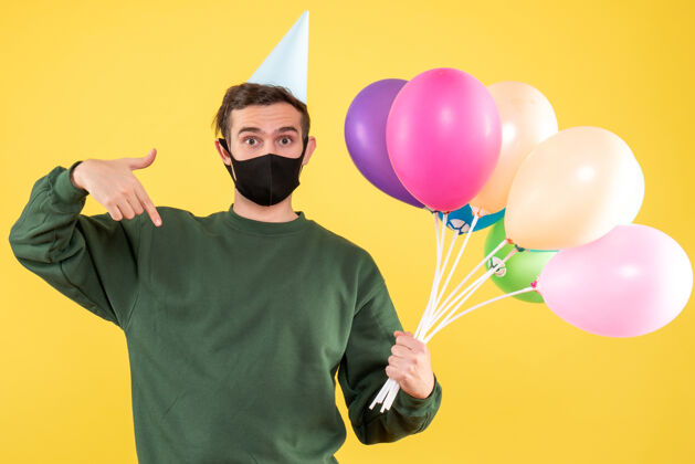 气球正面图：戴着派对帽的年轻人 手指朝下指着五颜六色的气球站在黄色的地面上生日聚会站着