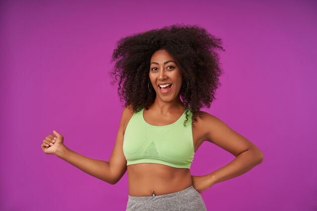 表情快乐的年轻卷曲的黑皮肤女士 穿着运动装 带着快乐的笑容 站在紫色的地面上 举起手 表现出积极的情绪牙齿卷发上衣