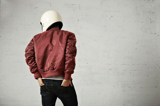 棒球服一个年轻的摩托车手 戴着白色头盔 穿着红色尼龙夹克 双手插在牛仔裤的后口袋里 从后面开枪射击 拍摄地点是一间有白色墙壁的工作室表情夹克飞行