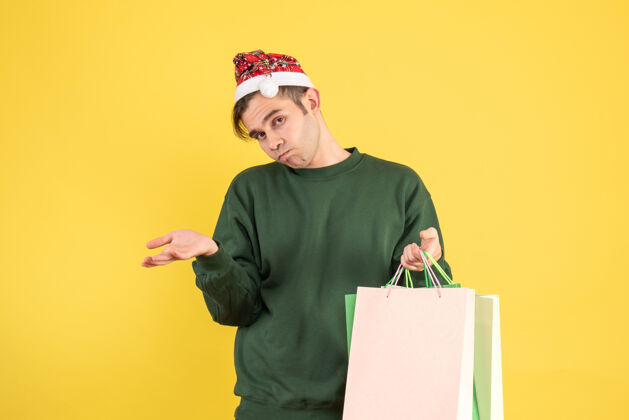 困惑的年轻人前视图困惑的年轻人拿着圣诞帽购物袋站在黄色抱着帽子站着