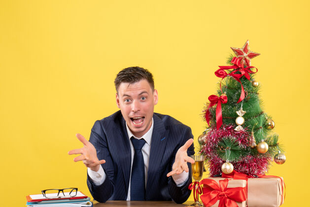 打开前视图中的快乐男人张开双手坐在圣诞树旁的桌子上 黄色的礼物坐着黄色快乐