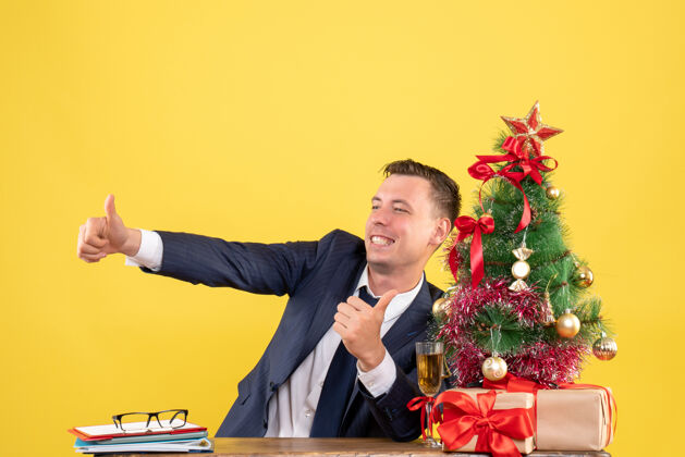 专业正面图是坐在圣诞树旁的快乐的年轻人竖起大拇指 黄色的礼物坐着黄色商人