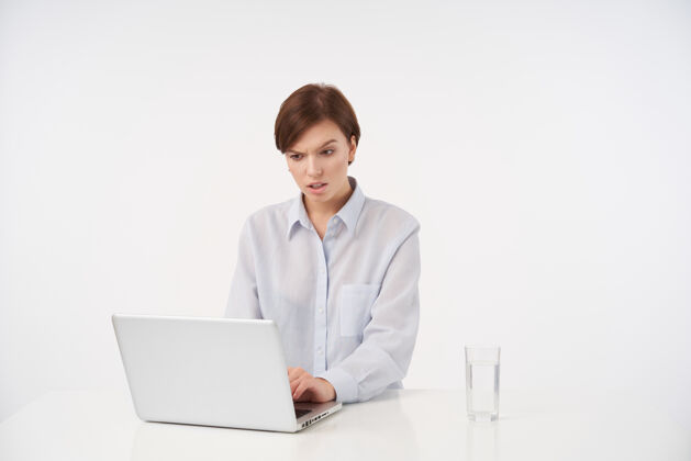 现代迷茫的年轻短发黑发女人 随意的发型 手放在笔记本电脑的键盘上 困惑地看着扬起眉毛的屏幕 孤立在白色上困惑化妆品员工