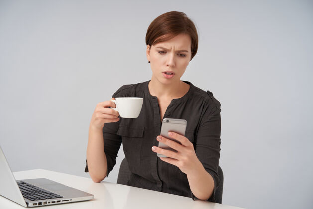脸困惑的棕色头发的年轻女士 短发时髦 眉头紧锁 一边看着手机屏幕 一边拿着一杯茶 一边摆着白色的姿势肖像短黑色