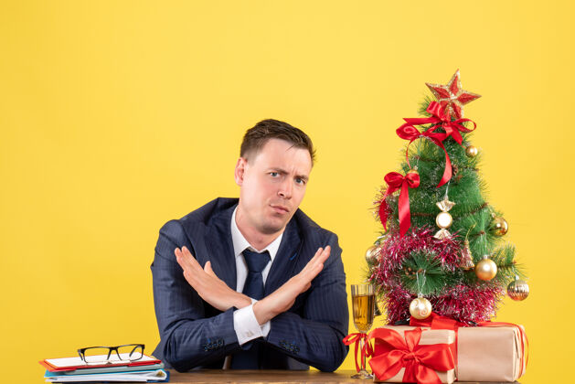 成人前视图：体贴的男人交叉双手坐在圣诞树旁的桌子旁 黄色的礼物商人桌子人