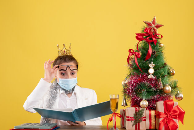 脖子正面图女医生戴着无菌口罩坐着 黄色背景上有文件 还有圣诞树和礼品盒成人文件面具