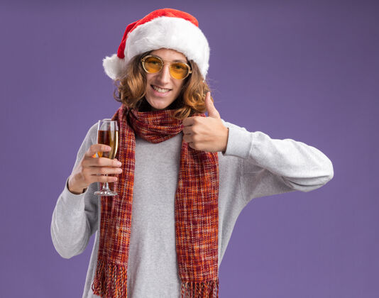 眼镜快乐的年轻人戴着圣诞老人的帽子 戴着黄色的眼镜 脖子上围着暖和的围巾 手里拿着一杯香槟 微笑着看着相机 竖起大拇指站在紫色的背景上背景圣诞老人展示