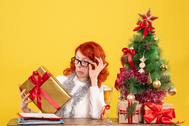 快乐前视图：女医生拿着圣诞礼物和黄色背景上的圣诞树坐着女医生前面医生