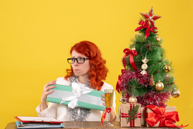 圣诞节前视图：女医生拿着圣诞礼物和黄色背景上的圣诞树坐着漂亮女性前面