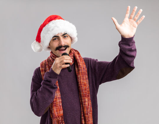 围着年轻的留着胡子的男人戴着圣诞老人帽 脖子上围着暖和的围巾 手里拿着麦克风 高高兴兴地微笑着 举起手臂站在白色的背景上抱着温暖站着