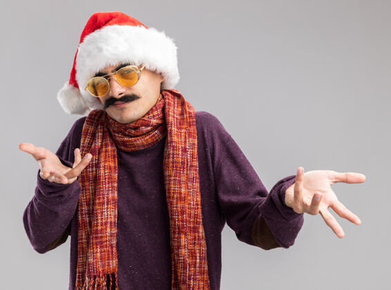 围巾一个留着胡子的男人戴着圣诞老人帽 戴着黄色眼镜 脖子上围着一条暖和的围巾 站在白色的背景上 困惑地看着镜头 伸出双臂快乐站着脖子