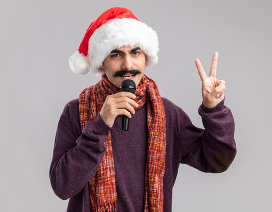 围着年轻的胡子男人戴着圣诞老人的帽子 脖子上围着暖和的围巾 对着麦克风说“v”字围巾圣诞老人圣诞