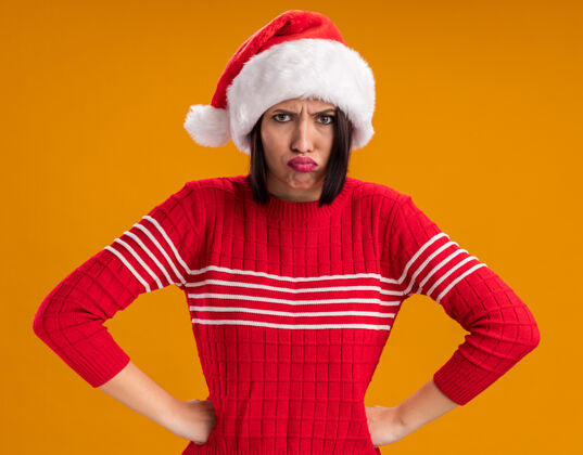 圣诞老人愤怒的年轻女孩戴着圣诞帽 手放在腰上 隔离在橙色的墙上愤怒穿年轻